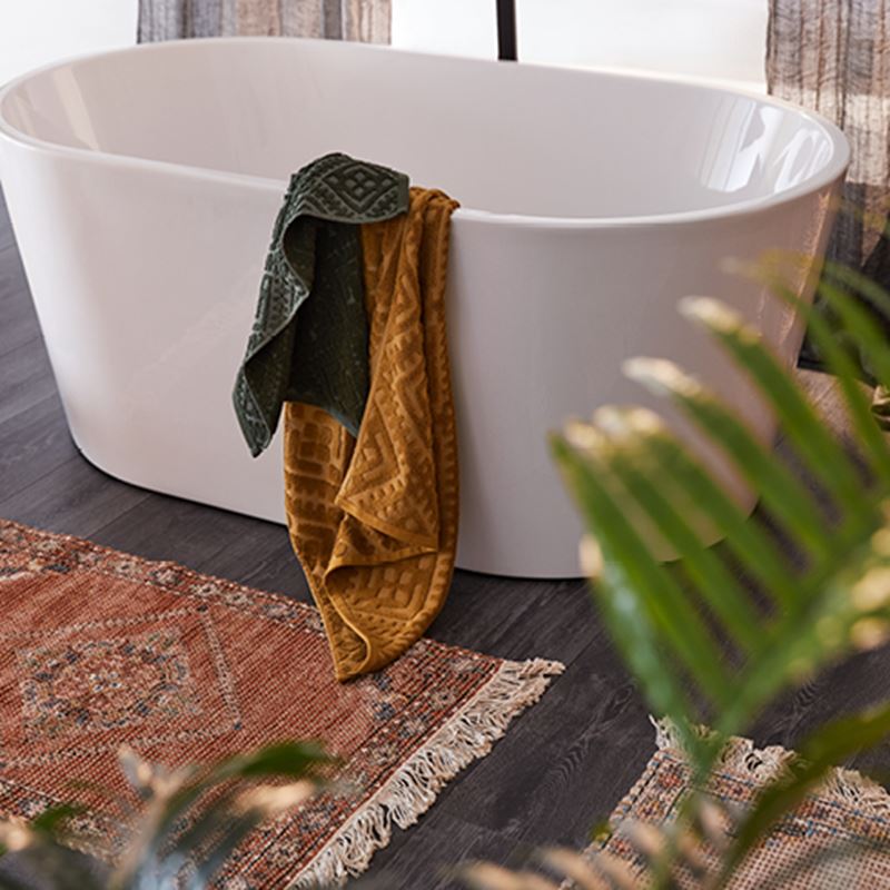 Free-standing bath with two textured towels hanging over the edge and a soft red Monte Mat with tasselled edging in front. 