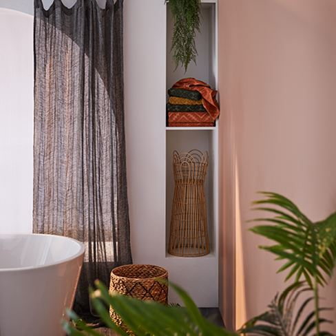 The corner of a bathroom featuring shelving with a plant stand, rattan basket, stack of warm-hued towels.