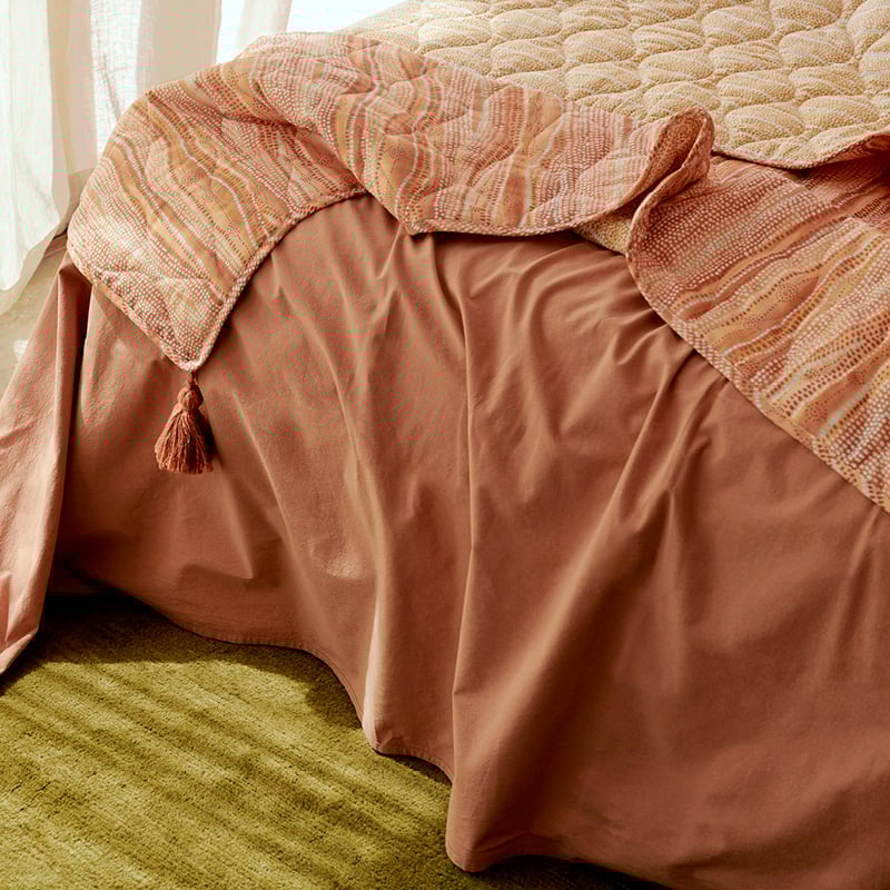 Solid earth coloured sheets styled on a bed, with a coverlet draped naturally over the top in contrasting lighter hues of orange, pink and yellow.