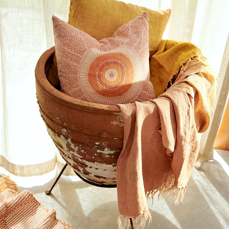 A rustic ceramic basket holds a cushion from the collection and draped linen throws, cushion has dotted design in red with bolder orange tones in centre.