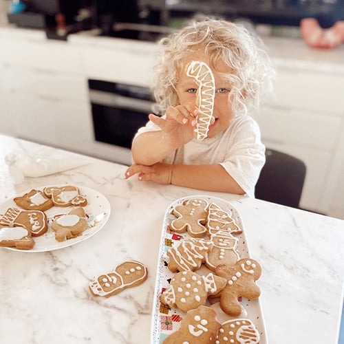 Toddler at kitchen bench holds a candy cane shaped ginger cookie in front of her face