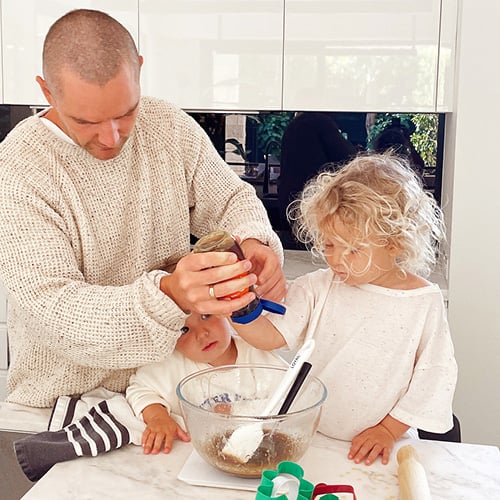 Father with two young children, baking Christmas gingerbread cookies, helps squeeze honey into a mixing bowl on a kitchen bench