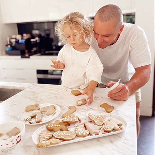 Father and toddler standing at a bench decorating Christmas gingerbread cookies.
