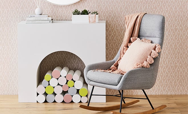 Pink cushion and quilt cover placed on a one-seater grey rocking chair. There is a white fire place in the background with pink toned walls and light brown floor boards