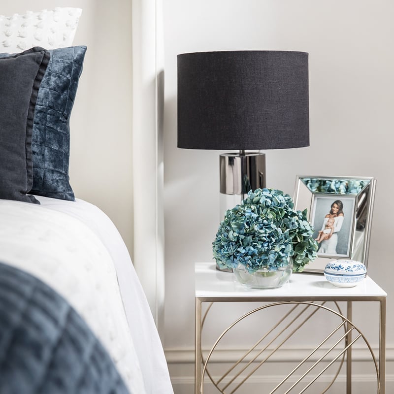 Brushed brass bedside table with marble top, with blue hued flower arrangement and mirror edged photo frame.