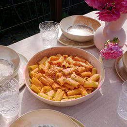 A bowl of pasta alla vodka on a table surrounded by serving ware, drinking glasses and pink vase filled with flowers.