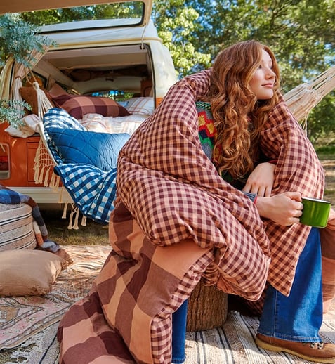 A woman sits in the back of an open campervan wrapped in a gingham quilt, rugs cover the ground beneath her feet and a knitted ottoman is visible in the left of the frame. Behind her hangs a hammock filled with cushions and a blue gingham blanket. 