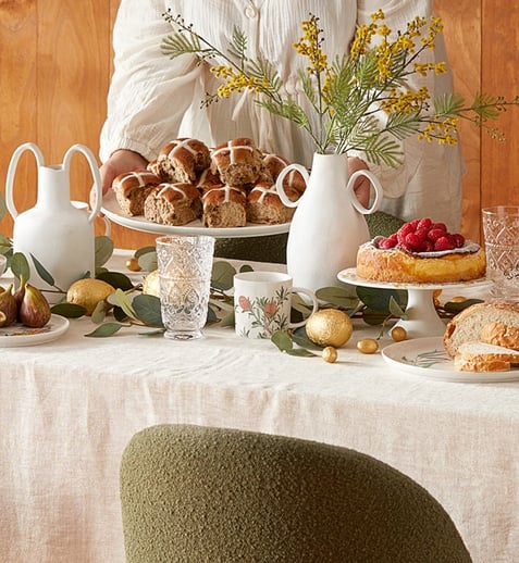 A dining room scene with Easter treats across the table, and green chairs. The background is a wooden wall, and the table is piled with serving ware, a linen tablecloth and fresh flowers.