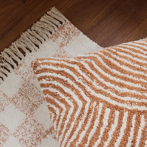 A close up shot of a textured white and brows stripe cushion, lying on a tasseled brown and white checked rug.