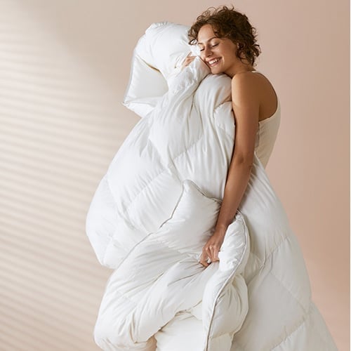 Woman/man standing cuddling a plush quilt pictured against a nude pink background. 