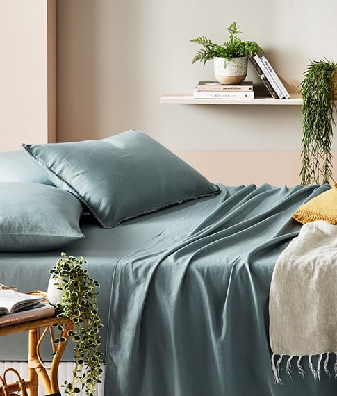 Side-on-view on Bamboo Linen Sage Sheets and matching pillowcases on a bed, next to a rattan side table with books. 