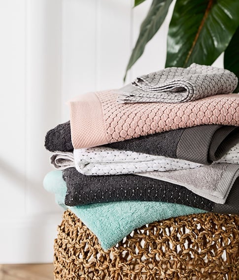 Stack of Bamboo Navara Towels including pink, coal, grey marle, white, and more – all on top of a rattan table. 