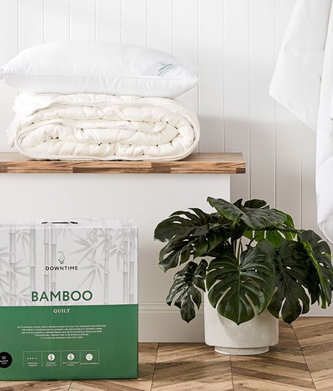 Bench seat with a quilt folded and a pillow on top, and in the foreground a plant next to a Downtime Bamboo Quilt in its box. 