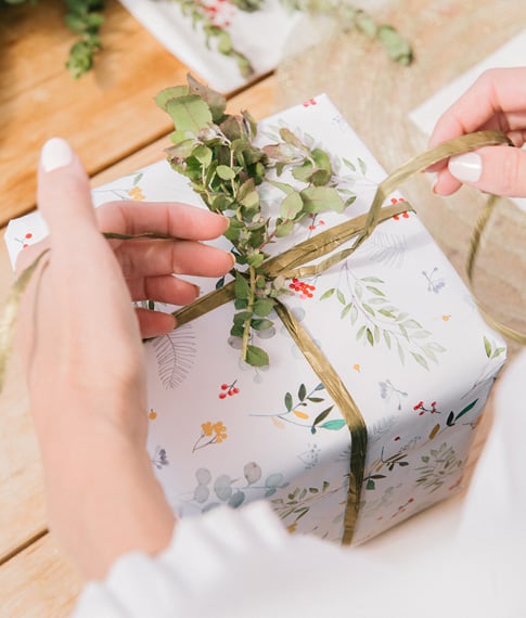 A woman/man adding the finishing touches to a Christmas present by with white paper and wrapping a green ribbon around the box.