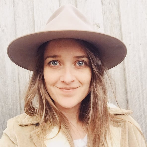 A close-up of a woman smiling wearing a natural beige coat and matching wide brimmed hat. 