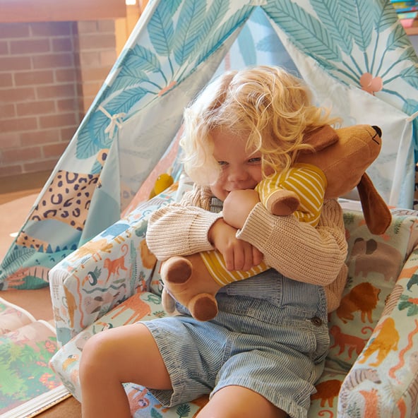 A child sits on a flip-out sofa cuddling a toy to the right of the frame, in front of a coloured teepee. There is a fire in a brick fireplace set in a brick wall in the background.