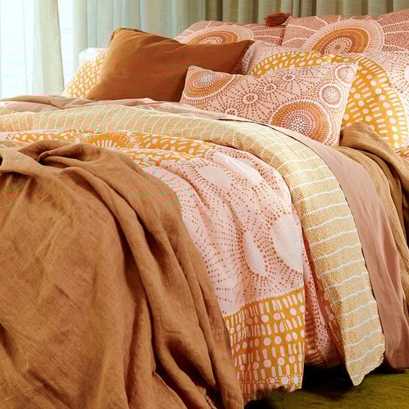 A styled bed in a focused shot displays the layered quilt cover in lighter shades of yellow, pink and orange while a rust linen throw and solid earth coloured sheets provide additional dimension. 