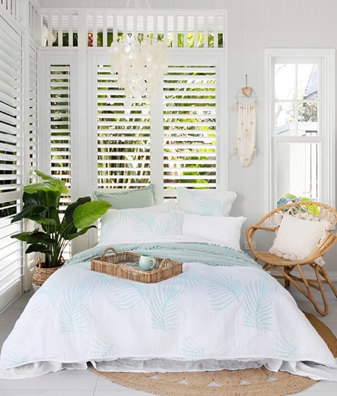 A front-on image of the coastal inspired bedroom with light blue and white bedlinen, rattan chair, tray, and pot plant.