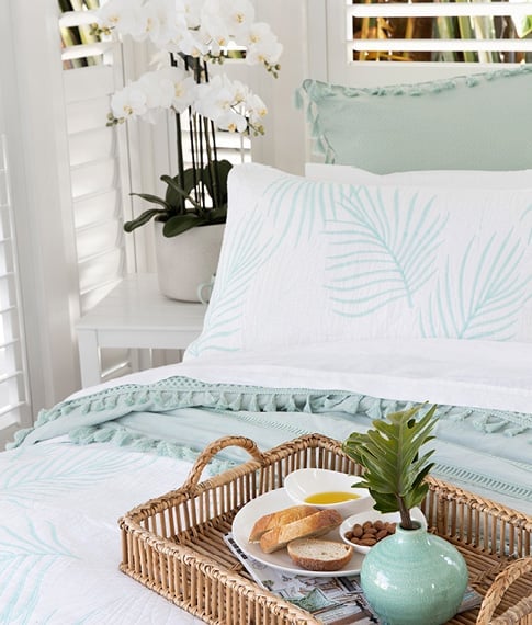 A rattan tray sits on top of a coastal inspired bed and in the background a beautiful white orchid frames the space.