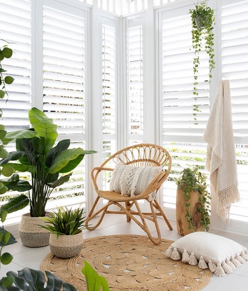 The corner of a room featuring white shutters, styled with a rattan chair, natural accessories and a multitude of plants.