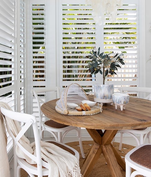 Corner of a room with white shutters, featuring a dining table with provincial chairs and styled with natural accessories.
