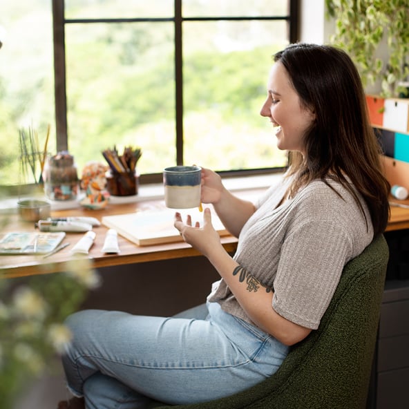 Claudia sits at her desk in her workspace having a coffee. The desk is surrounded by art and plants, and is in front of a window.