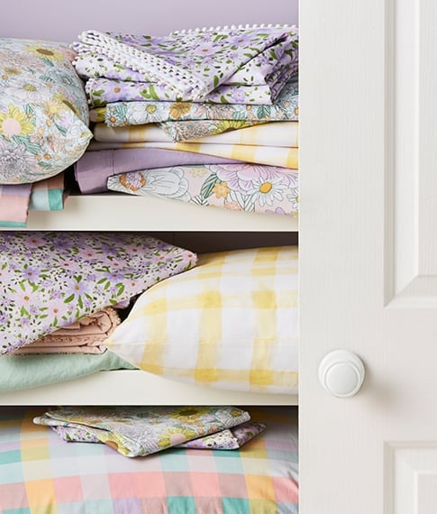 White linen cupboard partly open featuring bedlinen and pillows in pastels including plain, checks and floral patterns. 