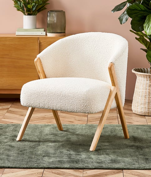 A white armchair in a boucle material sits in the foreground of a room, on an olive rug. 