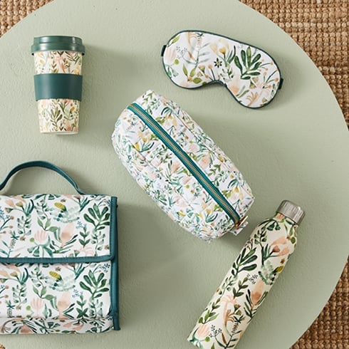 Table with accessories including drink bottle, reusable cup, eye mask and more on top – all with a botanical pink and green pattern. 