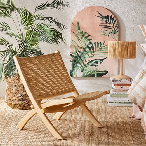 Burleigh Rattan Chair styled next to a bed with a rattan basket with Areca Palm, palm wall art and pile of books with bedside lamp on top. 