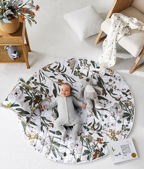 Baby laying on a nursery play mat with botanical print, and matching accessories including plush koala toy and cushion. 