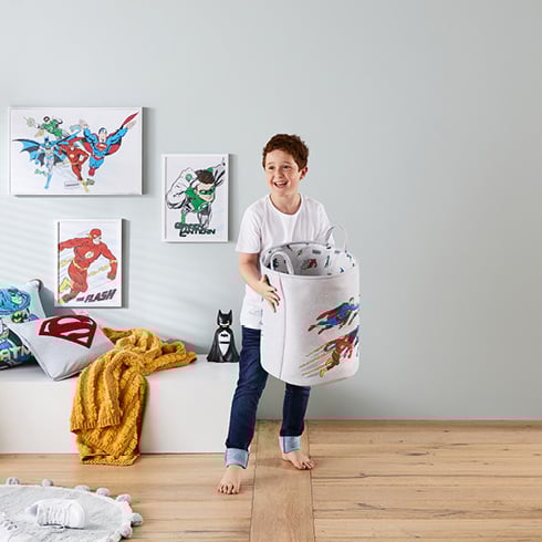 Boy holding Justice League Basket with matching wall art on the wall behind and cushions on a seating ledge. 