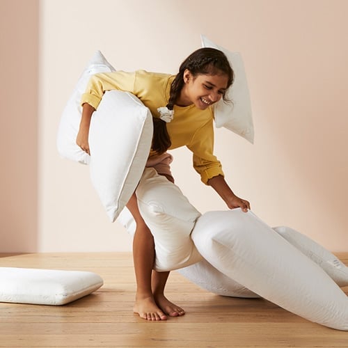 Child smiling standing holding a pillow and surrounded by pillows. 