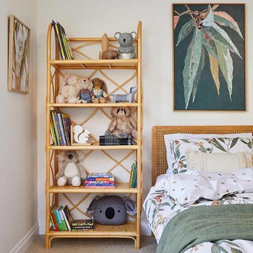 Corner of child’s bedroom next to bed is a rattan shelf featuring books and multiple plush toys.