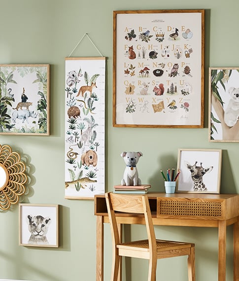A green wall featuring wall art in natural frames with a kid’s desk and matching rattan chair situated against the wall. 