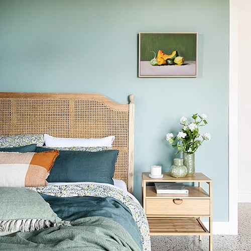 Bed styled with green hued bedlinen, matching cushion and throw on a bed with a rattan bedhead and a matching side table with one drawer. 