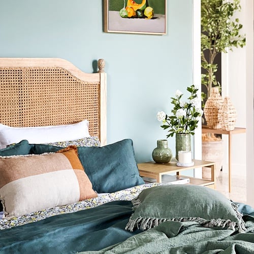 Angled view of a bed styled with green bedlinen, floral sheets and fringed cushion in orange and neutral colours. 