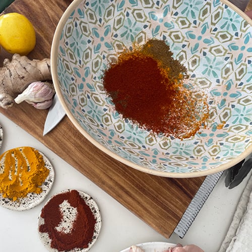 Bird’s eye view of kitchen bench with spices in a few bowls, and underneath the largest bowl is a chopping bord with fresh lemon, ginger, and garlic. 