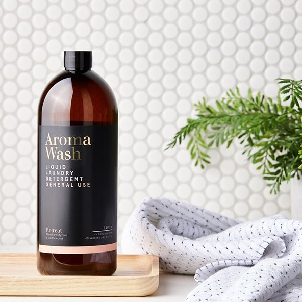 A bottle of Aroma Wash laundry liquid sits to the left of the frame, on a bamboo tray. To the left is a white towel with a black print, and a plant, all set in front of a tiled wall.