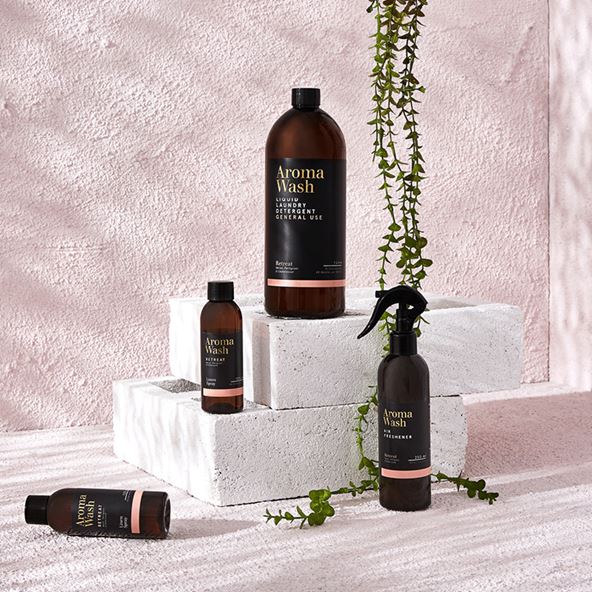 A stylised photo of a range of Aroma Wash products, arranged on white bricks with the tendrils of a plant above. The background is pink and textured.