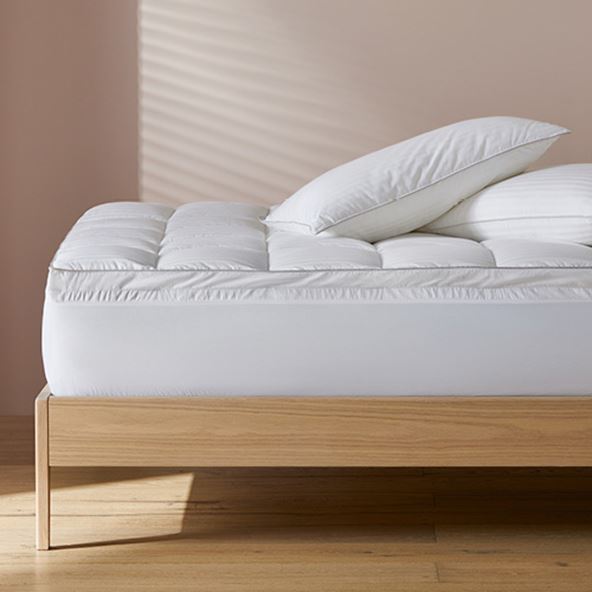 A side-on image of a bed featuring a mattress topper with two pillows stacked on top sitting against a neutral wall. 