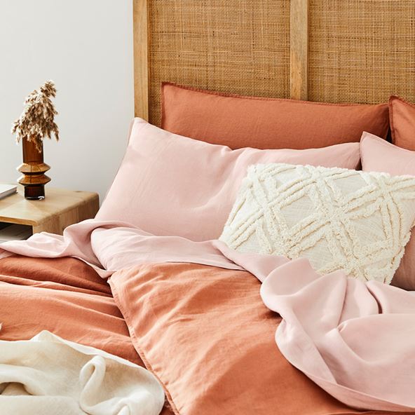 A close up shot of a bed with pale pink sheets and a rose-coloured quilt cover. A textured cushion decorates the bed and to the left in the background is a wooden side table with a geometric glass vase on top filled with dried stems.