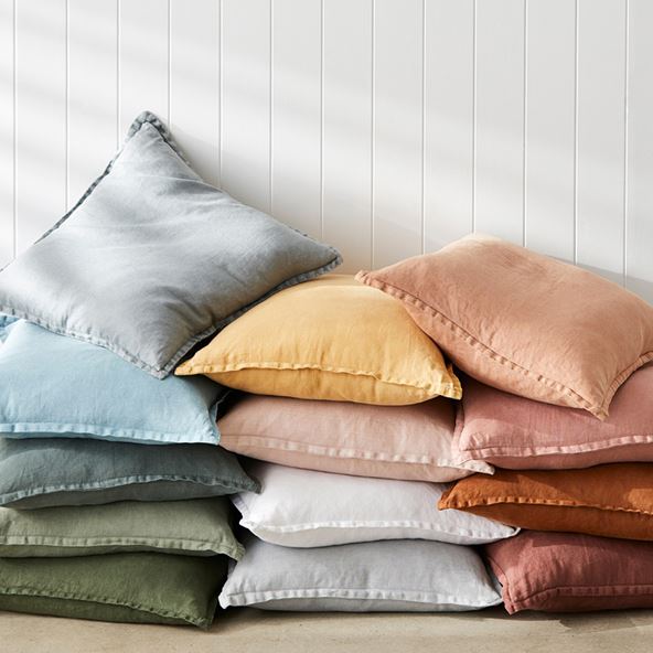A pile of 13 different coloured cushions is the focus of the shot, set off centre to the right of the photo. The background is a white, slatted wall and light from a window hits the pile of cushions.
