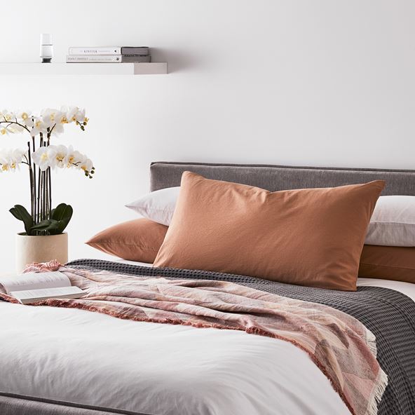 View of bed on an angle with white bedlinen, styled with warm toned accessories and a charcoal blanket. 