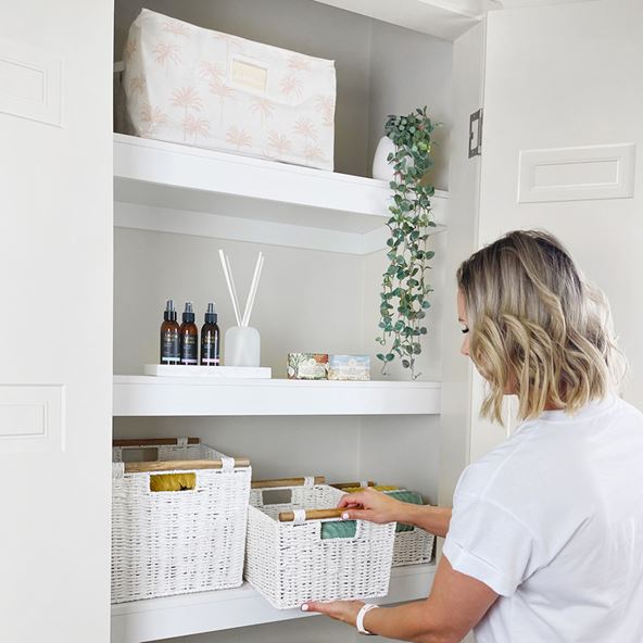 A woman places a white basket on the shelf of an organised cupboard.