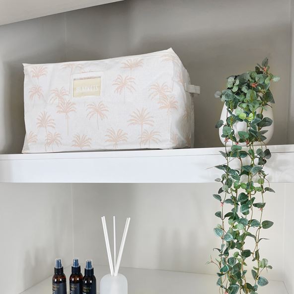 Large storage bag on a white shelf next to a potted green plant, above a shelf of linen sprays and a diffuser.