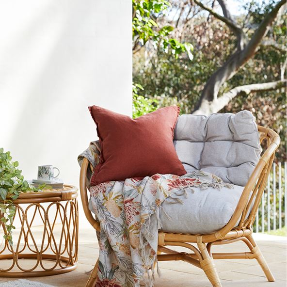 Outdoor scene with bamboo light brown coffee table and chair, dark orange and white cushions and floral throw
