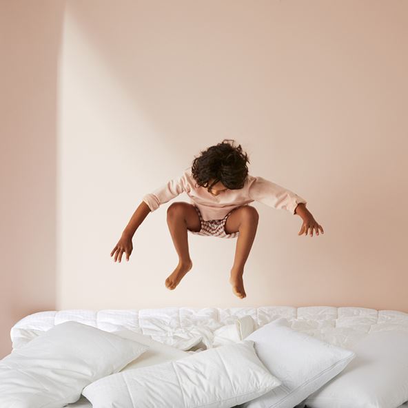 Child jumping mid-air on top of a bed made with a quilt on top and a pile of pillows below. 