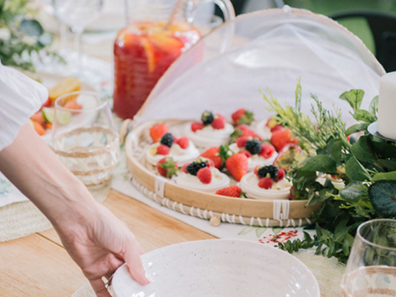 A woman/man setting the table with white tableware and in the background a natural rattan tray with mini pavlovas topped with berries. 