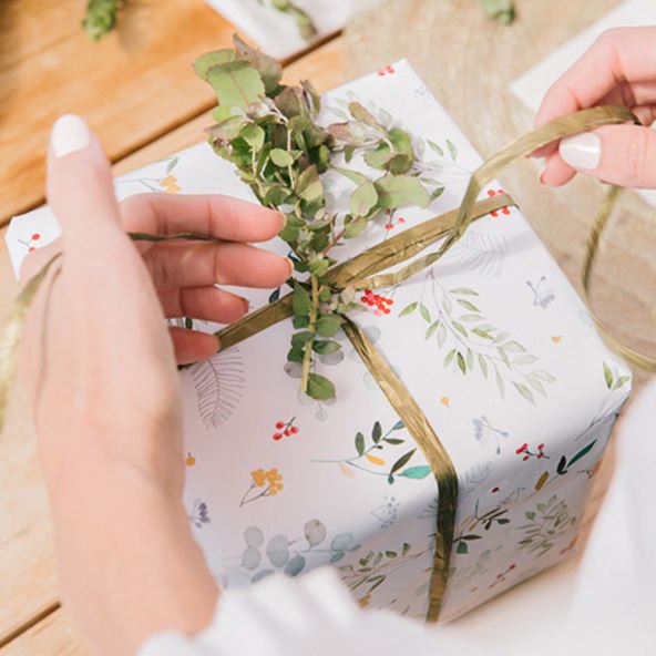 A woman/man adding the finishing touches to a Christmas present by with white paper and wrapping a green ribbon around the box.  